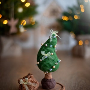 May include: A green crocheted Christmas tree ornament with white and blue beads and yellow ribbon. The tree is on a wooden base and there is a brown crocheted gift bag in front of it.