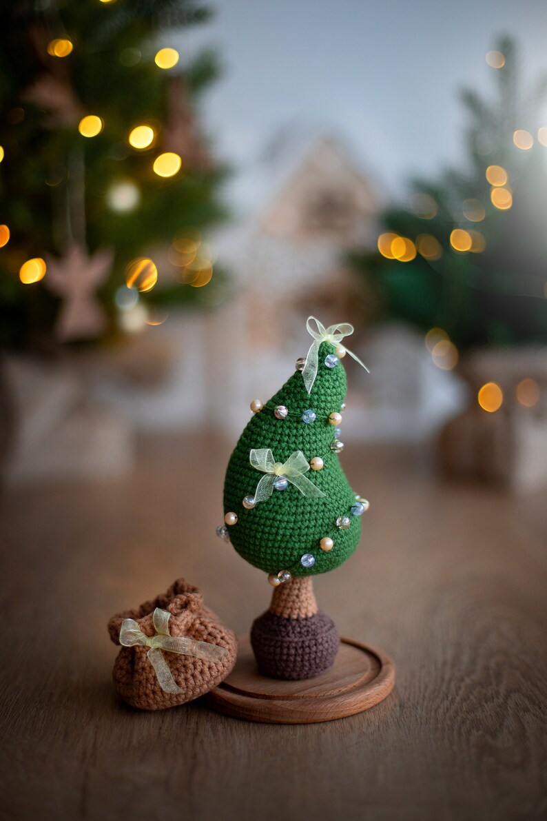 May include: A green crocheted Christmas tree ornament with white and blue beads and yellow ribbon. The tree is on a wooden base and there is a brown crocheted gift bag in front of it.