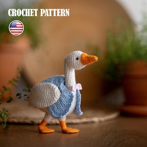 Op de afbeelding: Een gehaakte gans met een witte kop en lichaam, een oranje snavel en poten en een blauw jasje. De gans heeft een roze vlinderdas en staat op een houten oppervlak. De tekst "CROCHET PATTERN" staat bovenaan.