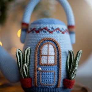 Pu&ograve; includere: Una teiera in crochet blu a forma di casa con una porta, finestre e due piante in vaso. La teiera ha un manico marrone e un bordo rosso e bianco.
