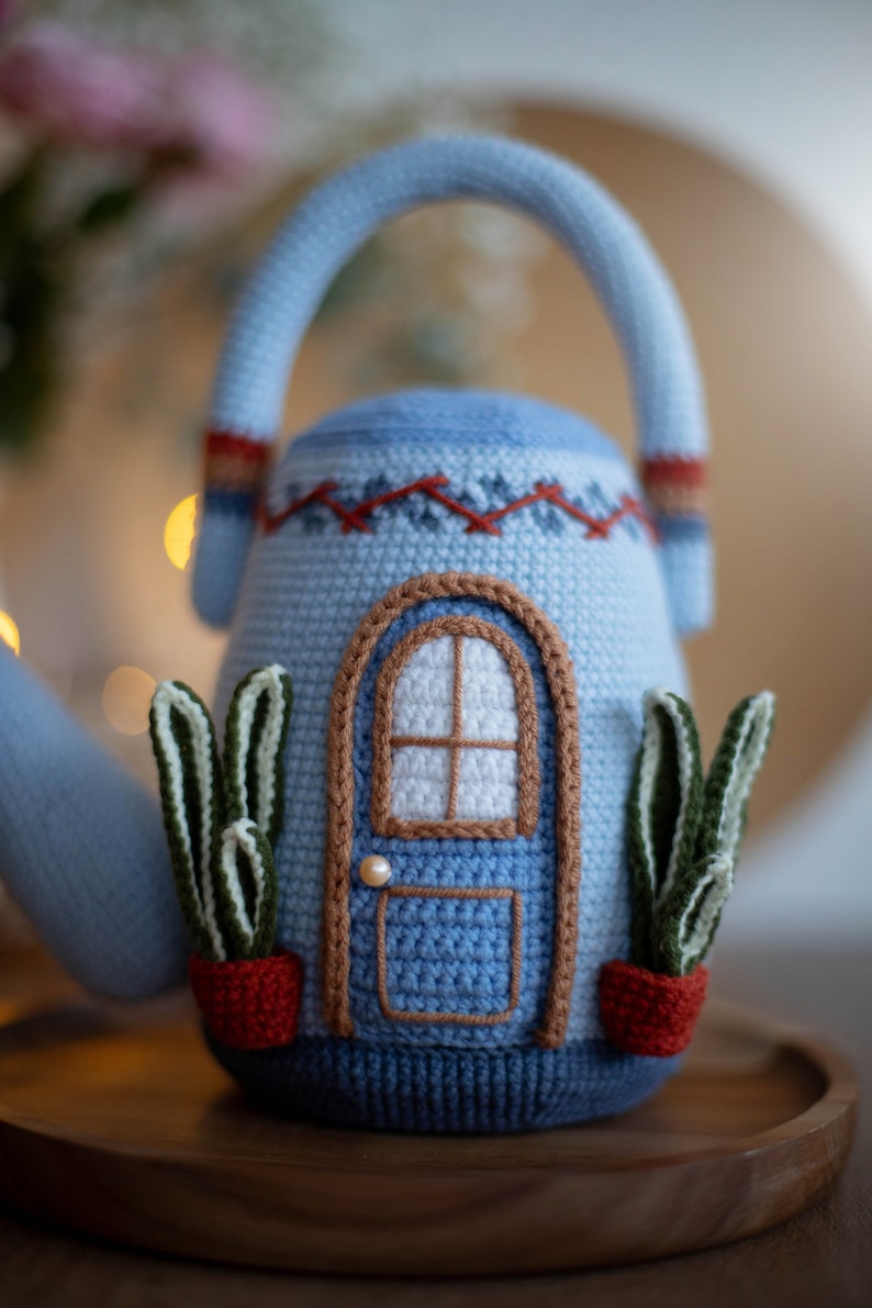 Pu&ograve; includere: Una teiera in crochet blu a forma di casa con una porta, finestre e due piante in vaso. La teiera ha un manico marrone e un bordo rosso e bianco.