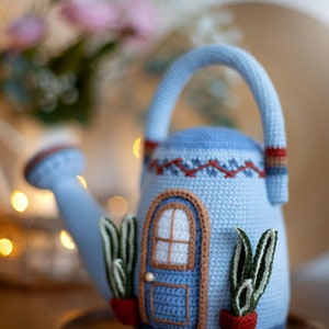 Pu&ograve; includere: Un annaffiatoio in crochet blu a forma di casa con una porta marrone e finestre bianche. L'annaffiatoio ha due piante verdi in vasi rossi sui lati.