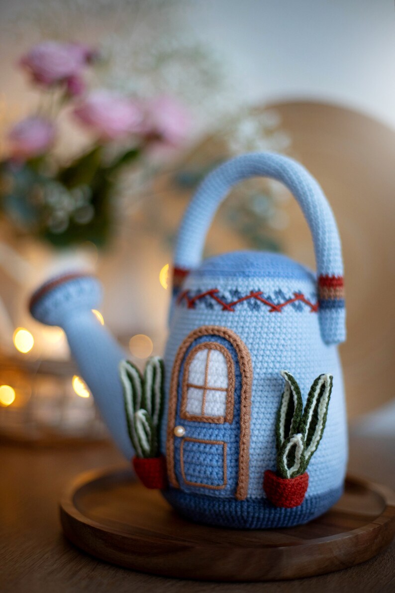 Pu&ograve; includere: Un annaffiatoio in crochet blu a forma di casa con una porta marrone e finestre bianche. L'annaffiatoio ha due piante verdi in vasi rossi sui lati.