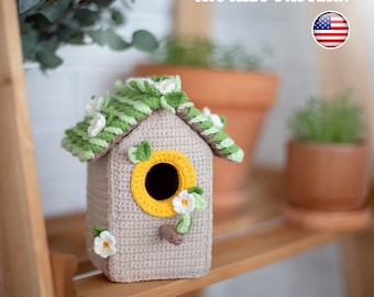 May include: Crochet birdhouse pattern. The birdhouse is beige with a green roof and yellow accents. It has a small opening for birds to enter. The birdhouse is decorated with white flowers.