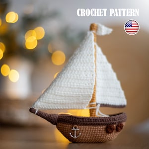 May include: Crochet pattern for a sailboat with a white sail and a brown hull. The sailboat has an anchor on the front and two small round details on the side. The pattern is available in US English.