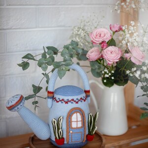Pu&ograve; includere: Un annaffiatoio blu all'uncinetto a forma di casa con una porta e finestre marroni. L'annaffiatoio &egrave; appoggiato su un vassoio di legno con un mazzo di rose rosa e fiori bianchi in un'anfora bianca dietro.