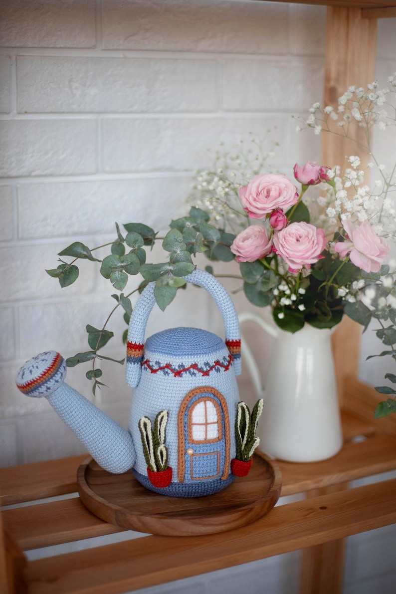 Pu&ograve; includere: Un annaffiatoio blu all'uncinetto a forma di casa con una porta e finestre marroni. L'annaffiatoio &egrave; appoggiato su un vassoio di legno con un mazzo di rose rosa e fiori bianchi in un'anfora bianca dietro.