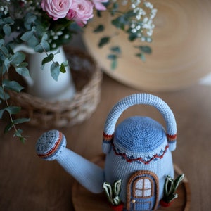 Pu&ograve; includere: Un annaffiatoio in crochet blu a forma di casa con una porta blu e una finestra. L'annaffiatoio &egrave; appoggiato su un vassoio di legno con piante verdi e carote che spuntano dai lati.