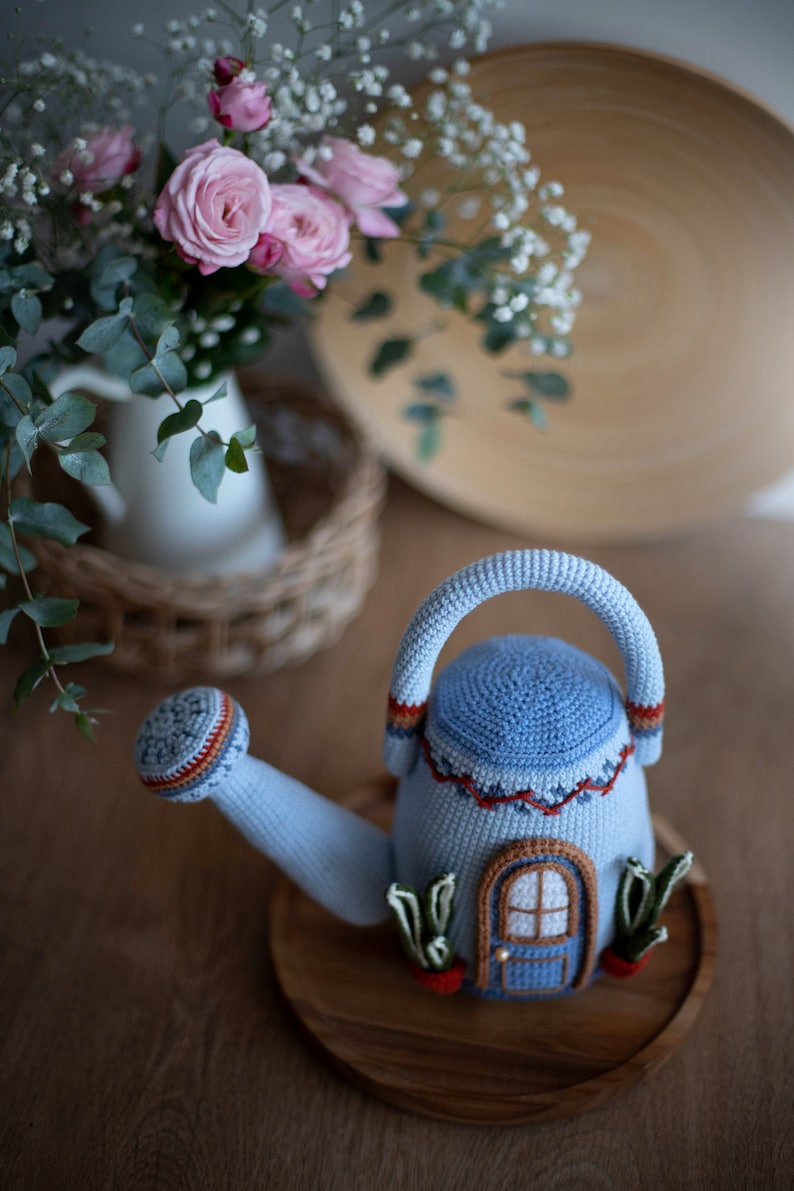 Pu&ograve; includere: Un annaffiatoio in crochet blu a forma di casa con una porta blu e una finestra. L'annaffiatoio &egrave; appoggiato su un vassoio di legno con piante verdi e carote che spuntano dai lati.