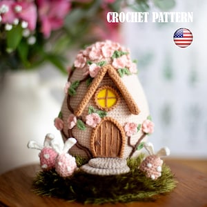 Può includere: Modello a uncinetto per una casa di uova di Pasqua fantasiosa con una porta rosa e marrone, una finestra gialla e una base verde. La casa è decorata con fiori rosa e foglie verdi. Il modello è disponibile in inglese.