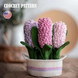 May include: Crocheted hyacinth flowers in a white basket with green leaves. The flowers are in shades of pink and purple. The basket has a striped design. The text "CROCHET PATTERN" is at the top of the image.