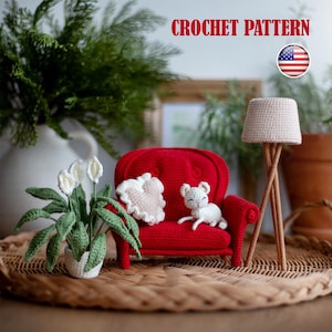 May include: Crochet pattern for a red armchair with a white crocheted pillow and a white crocheted cat sitting on it. The armchair is on a wicker surface with a white crocheted lamp in the background.