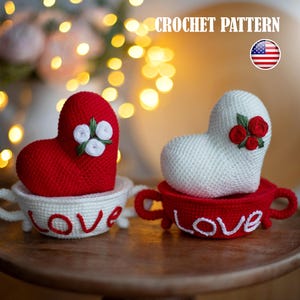 May include: Two crocheted heart-shaped ornaments with white and red yarn. Each heart is sitting in a crocheted cup with the word "LOVE" written on the front. The hearts are decorated with crocheted flowers.