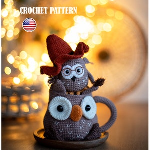 May include: A crocheted owl wearing glasses and a red hat sits inside a crocheted owl mug. The owl mug has a brown body, white eyes, and an orange beak. The owl mug is sitting on a wooden coaster.