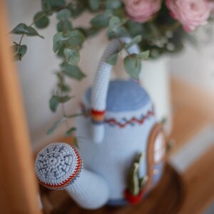 Pu&ograve; includere: Un annaffiatoio in crochet azzurro con un bordo rosso e arancione. L'annaffiatoio ha un motivo a fiocco di neve sul beccuccio e un piccolo cactus sul lato.