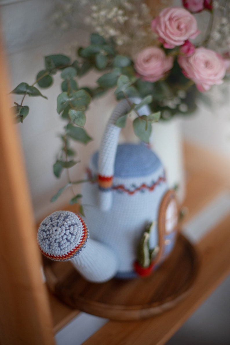 Pu&ograve; includere: Un annaffiatoio in crochet azzurro con un bordo rosso e arancione. L'annaffiatoio ha un motivo a fiocco di neve sul beccuccio e un piccolo cactus sul lato.