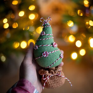 May include: A green crocheted Christmas tree ornament with red and white twine wrapped around it. The tree has small red and white rosettes for ornaments. The tree is sitting on top of a brown crocheted bag with red and white twine tied around it.