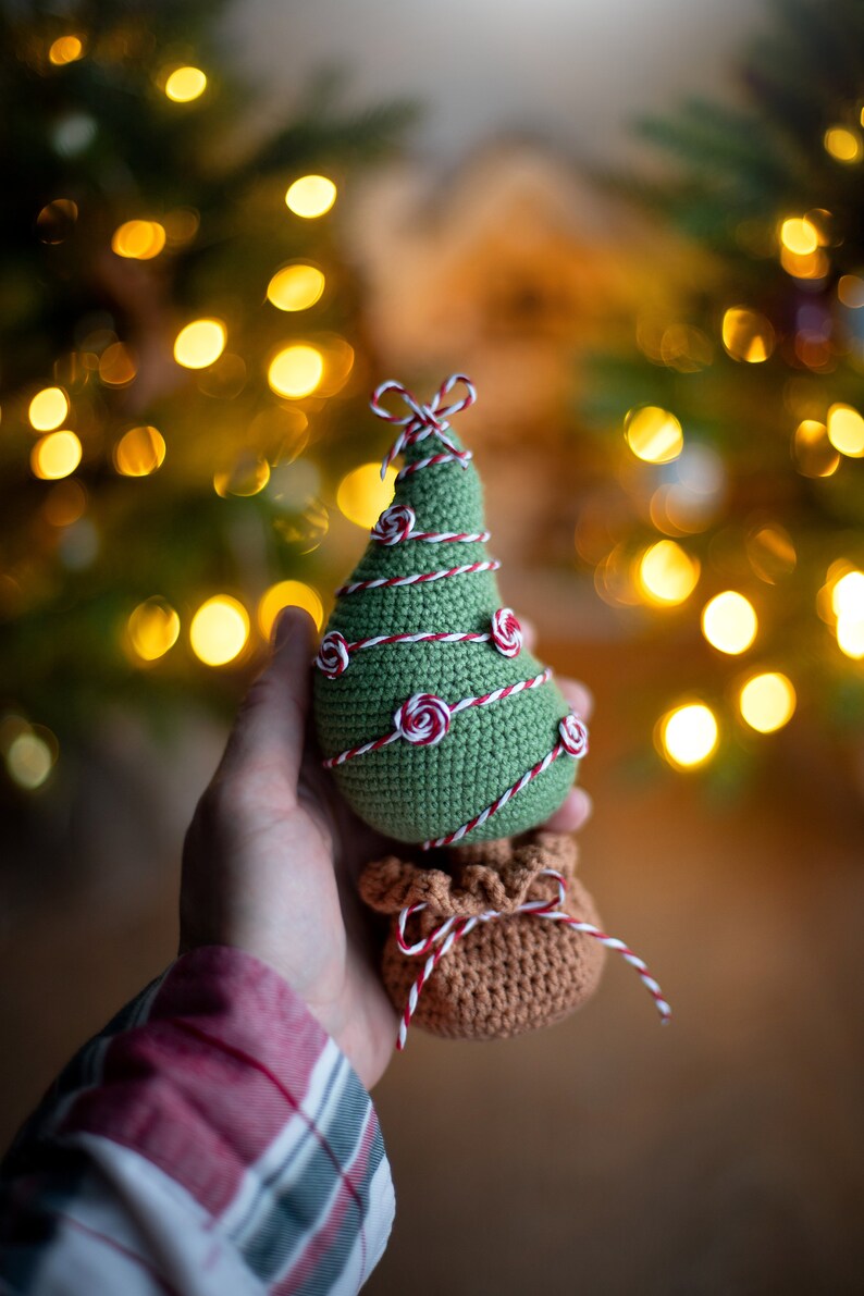 May include: A green crocheted Christmas tree ornament with red and white twine wrapped around it. The tree has small red and white rosettes for ornaments. The tree is sitting on top of a brown crocheted bag with red and white twine tied around it.