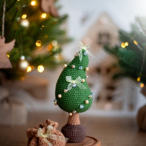 May include: A green crocheted Christmas tree ornament with white beads and yellow bows, standing on a wooden base. A brown crocheted gift bag with a yellow bow sits in front of the tree.