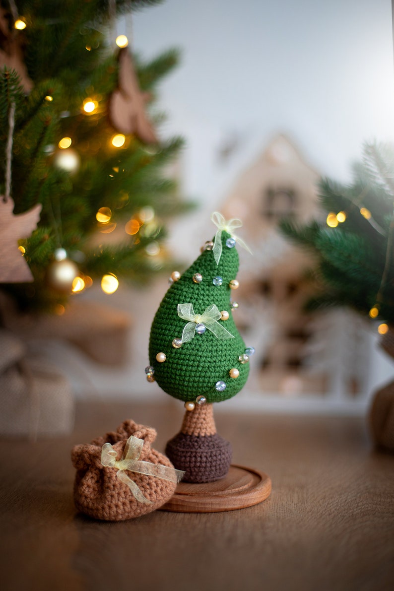 May include: A green crocheted Christmas tree ornament with white beads and yellow bows, standing on a wooden base. A brown crocheted gift bag with a yellow bow sits in front of the tree.