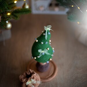May include: A green crocheted Christmas tree with white and blue beads and yellow bows on a wooden base. A brown crocheted bag with a yellow bow sits in front of the tree.