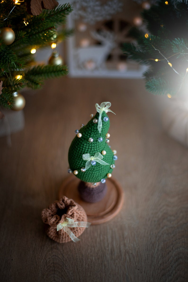 May include: A green crocheted Christmas tree with white and blue beads and yellow bows on a wooden base. A brown crocheted bag with a yellow bow sits in front of the tree.