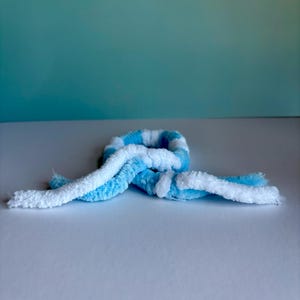 May include: A handmade, plush scarf in a looped design. The scarf features alternating stripes of white and light blue. The soft, textured material is visible in the close-up shot, highlighting the cozy nature of the accessory.