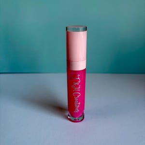Könnte beinhalten: Eine Tube leuchtend pinkfarbenen Lipgloss mit hellrosa Kappe und dem silbernen Text "T & H Creations". Die transparente Tube zeigt die leuchtende Farbe des Glanzes. Der Hintergrund ist einfarbig türkis.