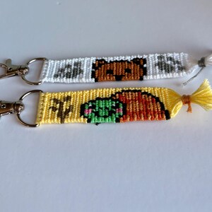May include: Two handmade keychains with silver clasps. One is white with a brown bear design, the other is yellow with a green and brown design. Both have decorative tassels. These keychains are perfect for adding a personal touch to your keys or bag.