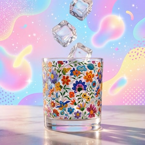 May include: A clear glass tumbler featuring a vibrant, multi-coloured floral pattern with orange, blue, purple, and yellow blossoms. Three clear ice cubes are suspended in the air above the glass, which sits on a smooth, reflective surface.