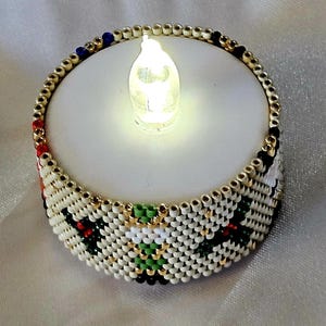 Könnte beinhalten: Eine batteriebetriebene LED-Kerze mit Perlendesign. Die weiße Kerze hat einen goldperlenbesetzten Rand und ein weißes und goldperlenbesetztes Design um die Basis, mit Akzenten aus roten, grünen und schwarzen Perlen. Die Kerze ist beleuchtet und strahlt sanft.