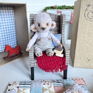 Könnte beinhalten: Ein handgefertigter Teddybär-Puppe in grauen Latzhosen sitzt in einem Miniatur-Sessel mit Hahnentrittmuster und einem roten Fransenüberwurf. Kleine Bücher und ein Schaukelpferd deuten auf eine gemütliche Szene hin.