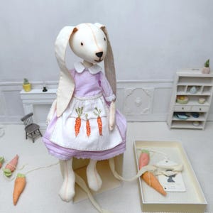 Pode incluir: Uma boneca de coelho de tecido feita à mão com orelhas longas, vestindo um vestido roxo claro e um avental branco bordado com três cenouras laranja. A boneca tem um rosto costurado e está posicionada perto de uma caixa com duas cenouras de tecido.