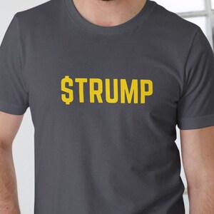 May include: A dark gray t-shirt with the word "$TRUMP" printed in yellow. The text is in a pixelated font. The shirt is short-sleeved and has a crew neck.