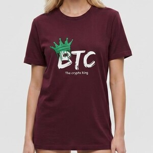 May include: A maroon short-sleeved t-shirt featuring a graphic print on the chest. The design includes a green crown sitting atop the white text "BTC" and the smaller white text "The crypto king" printed directly underneath the main letters.