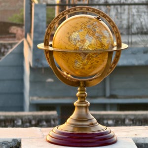 May include: A vintage-style globe with a golden finish, mounted on a brass stand with a dark red base. The globe features detailed geographical markings and is encircled by a brass meridian ring with degree markings. A decorative and educational item.