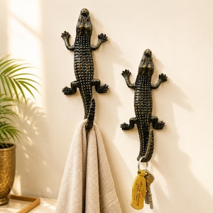 May include: Two wall-mounted hooks shaped like bronze-colored crocodiles with textured scales. The left hook holds a beige towel, while the right hook supports a metal keyring with a yellow tag featuring black text in a non-Latin script.