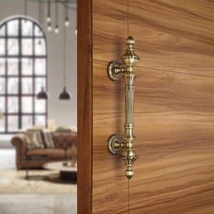 May include: Ornate brass door pull handle with a vertical, ribbed design. The handle features decorative finials at the top and bottom, and circular backplates. The door is made of wood with a natural grain.