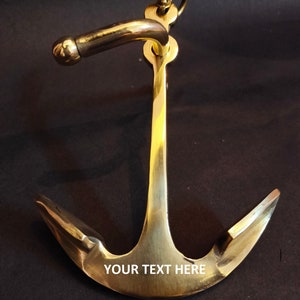 Può includere: Un'ancora in ottone con un manico curvo e un anello in alto. L'ancora ha un design classico con il testo "YOUR TEXT HERE" sul davanti. L'ancora è un oggetto decorativo nautico.