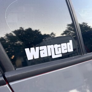 May include: A white "Wanted" sticker is affixed to a car window. The sticker's bold, sans-serif font stands out against the dark background of the window and the gray car door. The sticker is centered on the window.