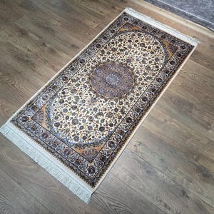 Könnte beinhalten: Ein rechteckiger Teppich mit einem komplizierten Blumenmuster in Creme-, Marineblau- und Goldbrauntönen. Der Teppich hat ein zentrales kreisförmiges Medaillon, eine dekorative Bordüre und weiße Fransen an den beiden kürzeren Enden des Stücks.