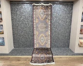 May include: A long, rectangular rug with a detailed, symmetrical pattern. The rug features a central design with repeating oval and circular motifs in shades of gold, brown, and cream, bordered by a deep burgundy. The rug has a fringed edge.