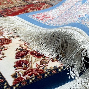 May include: Close-up of a rolled-up area rug with a floral pattern. The rug features a cream-colored field with red roses and green leaves. A blue border frames the design, and the rug has white fringe.