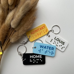 Puede incluir: Cuatro llaveros con etiquetas de diferentes colores. Las etiquetas tienen letras en braille y las palabras "KONTAKT", "WATER", "LOUIS" y "HOME".
