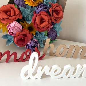 Peut inclure: Un bouquet de fleurs en papier colorées, rouges, jaunes, roses et bleues. Les mots décoratifs en bois "amor", "home" et "dream" sont au premier plan. Les mots sont rouges, beiges et blancs.