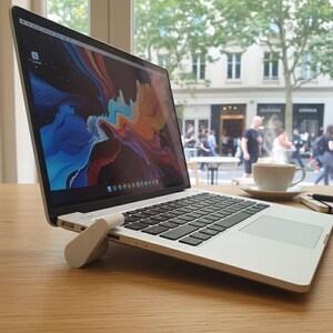 Puede incluir: Un ordenador portátil plateado con un teclado negro y una pantalla abstracta colorida está sobre una mesa de madera. Una taza y un platillo blancos, un cuaderno y un bolígrafo están al lado del portátil. Un soporte blanco para portátil está adjunto en la parte inferior.