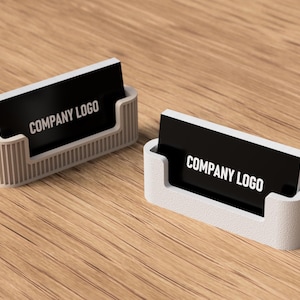 Pode incluir: Dois porta-cartões de visita com cartões pretos exibindo "COMPANY LOGO" em texto branco. Os porta-cartões são cinza claro e têm formato retangular com uma ranhura para guardar os cartões. O fundo é uma superfície de madeira.