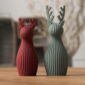 Puede incluir: Dos figuras decorativas de ciervos. Una es roja, la otra verde. Ambas tienen un diseño de rayas verticales y astas. Están colocadas sobre una superficie de madera. El fondo está borroso.