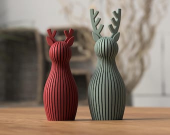 Può includere: Due statuette decorative a forma di cervo. Una è rossa, l'altra verde. Entrambe hanno un design a righe verticali e corna. Sono posizionate su una superficie di legno. Lo sfondo è sfocato.