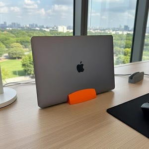 Puede incluir: Un ordenador portátil gris con el logotipo de Apple está apoyado por un soporte naranja sobre un escritorio de madera clara. Una alfombrilla de ratón negra y un soporte gris también están en el escritorio. El fondo muestra un paisaje urbano y un cielo azul.
